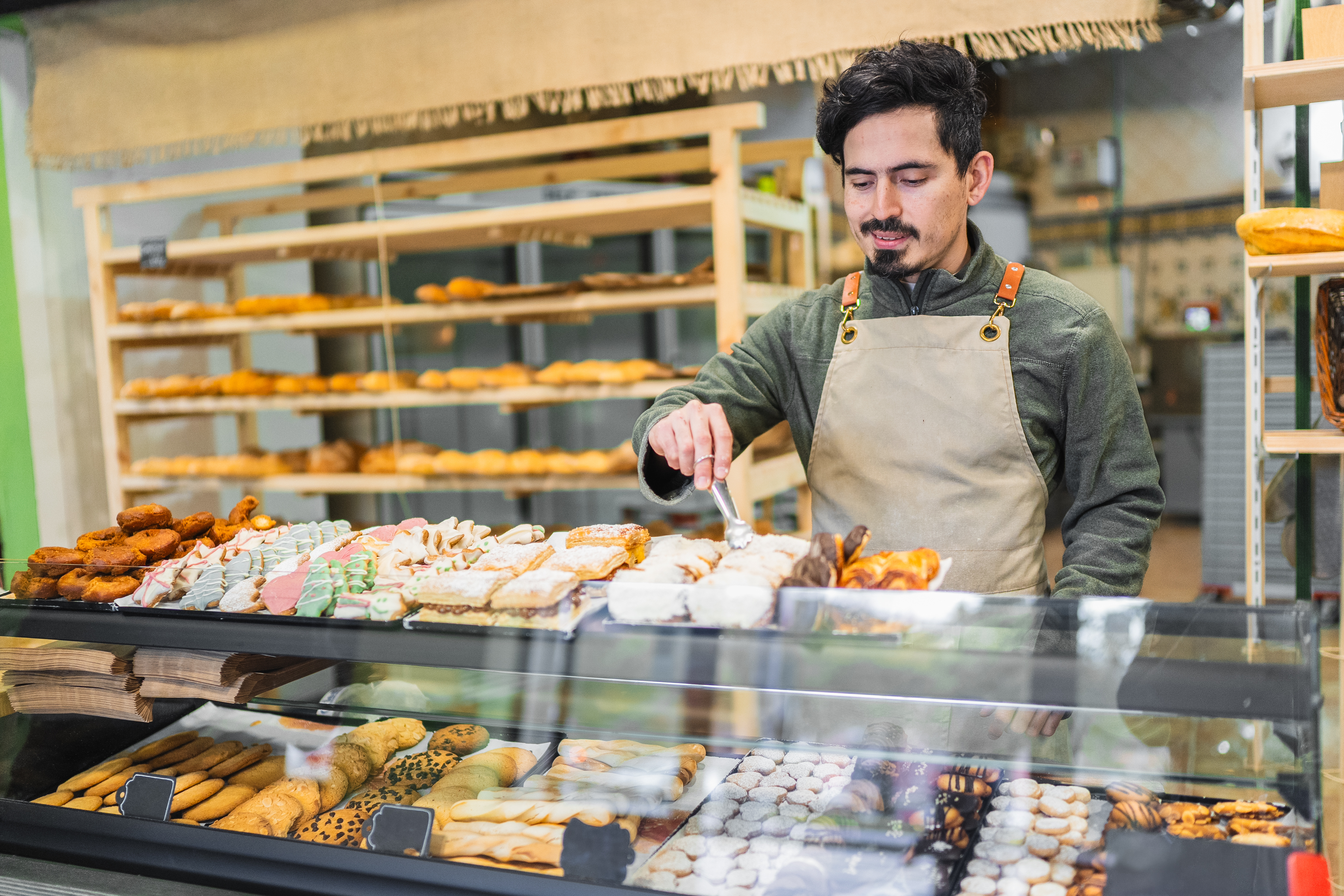Baker using tongs to arrange delicious pastries in a display case at a bakery shop, showcasing a variety of baked goods